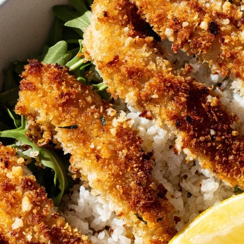 A close-up of the Crispy Parmesan Chicken Cutlet Bowl highlights crunchy, golden-brown cutlets layered on fluffy rice with vibrant greens.  